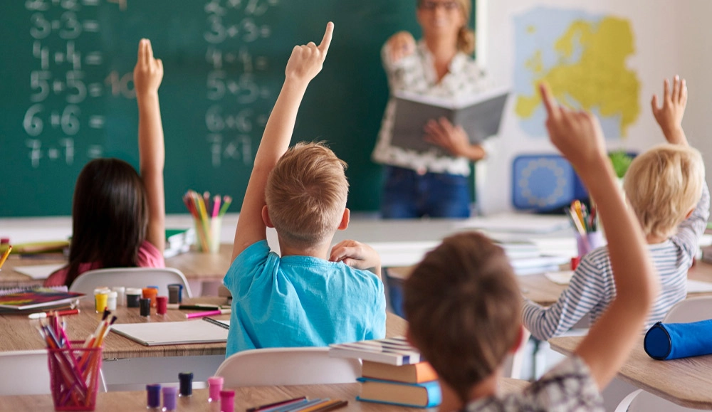 Crianças com o braço levantado durante aula para responder algum questionamento da professora.